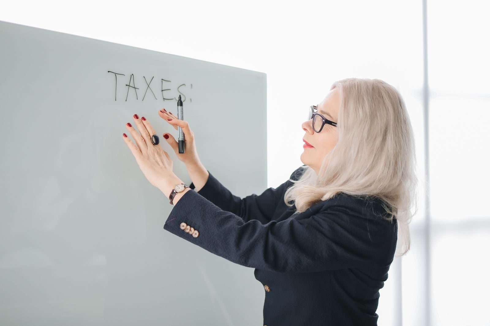 Elderly woman in a black blazer writes 'TAXES' on a whiteboard indoors