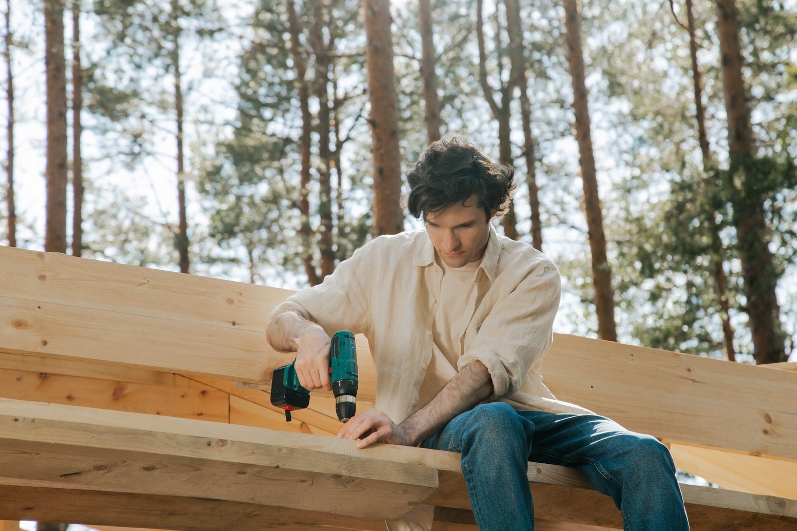 Focused carpenter uses power drill on wooden beams in an outdoor setting, surrounded by trees