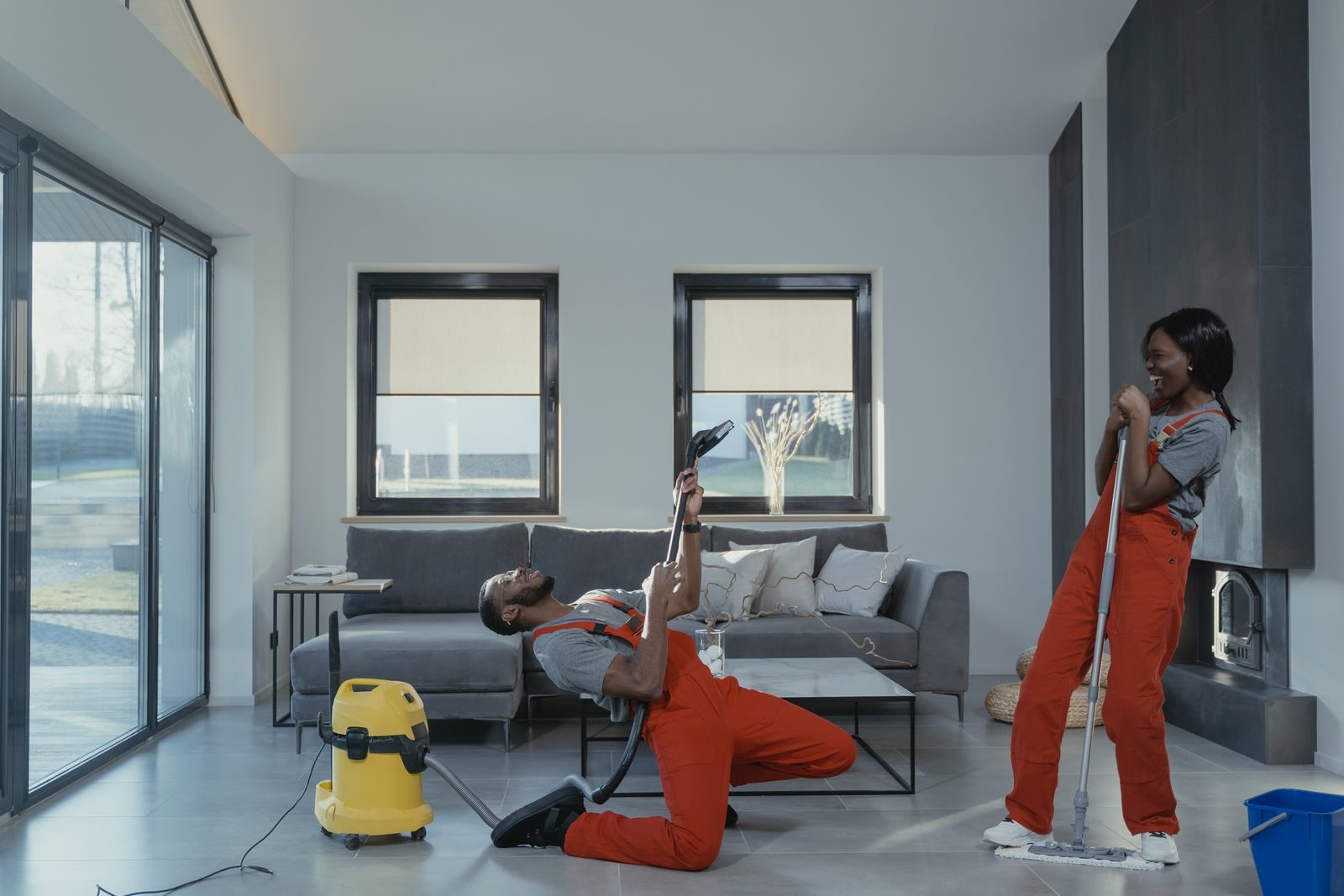 Fun and engaging cleaning session with a man and woman in overalls using cleaning tools in a modern living roo