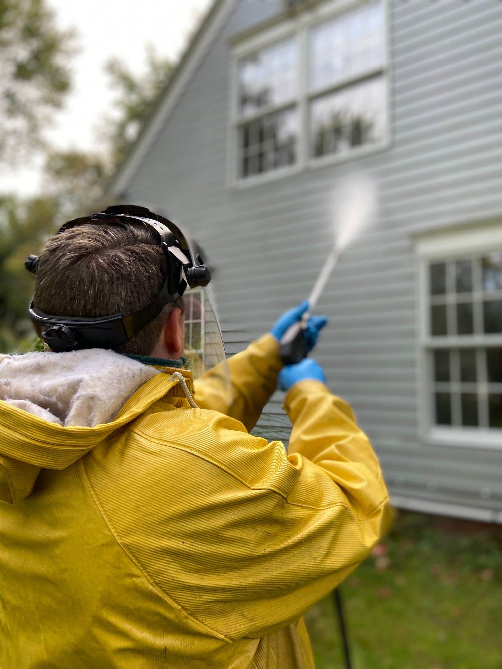Person cleaning house siding with a pressure washer, wearing protective gear in a yellow raincoat