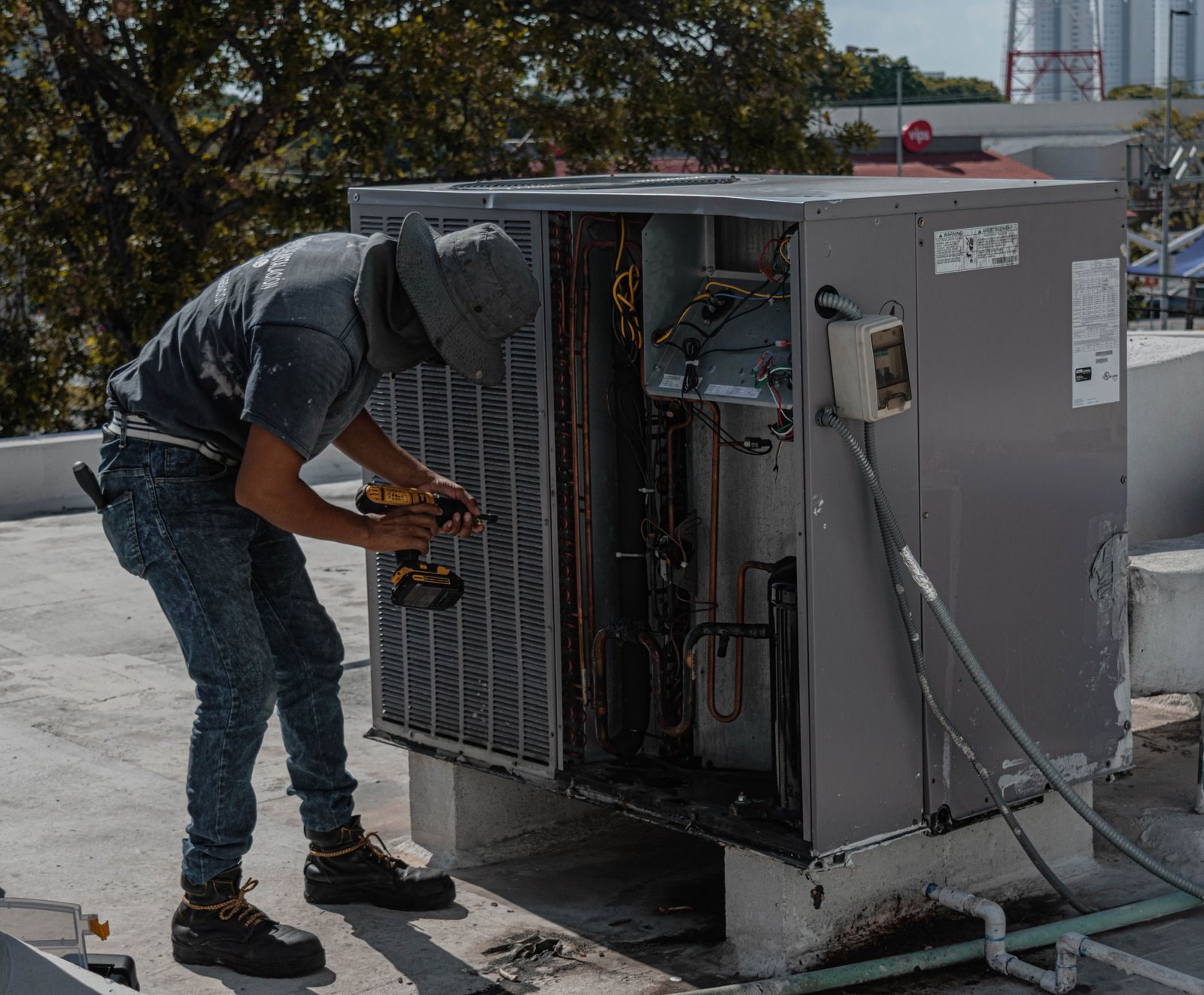 A worker in a bucket hat repairs an outdoor air conditioning unit on a rooftop