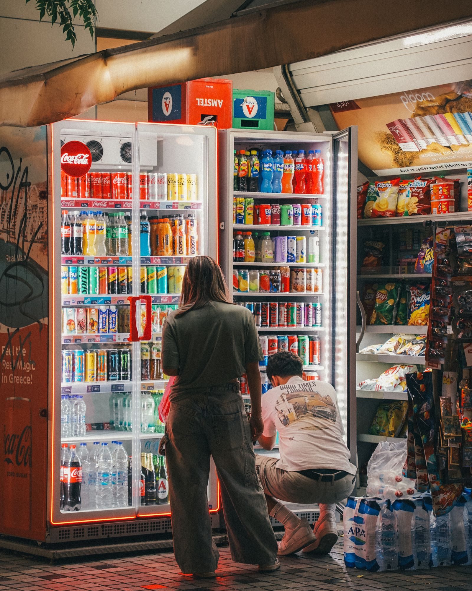 People selecting drinks at a convenience store aisle illuminated by vending machines