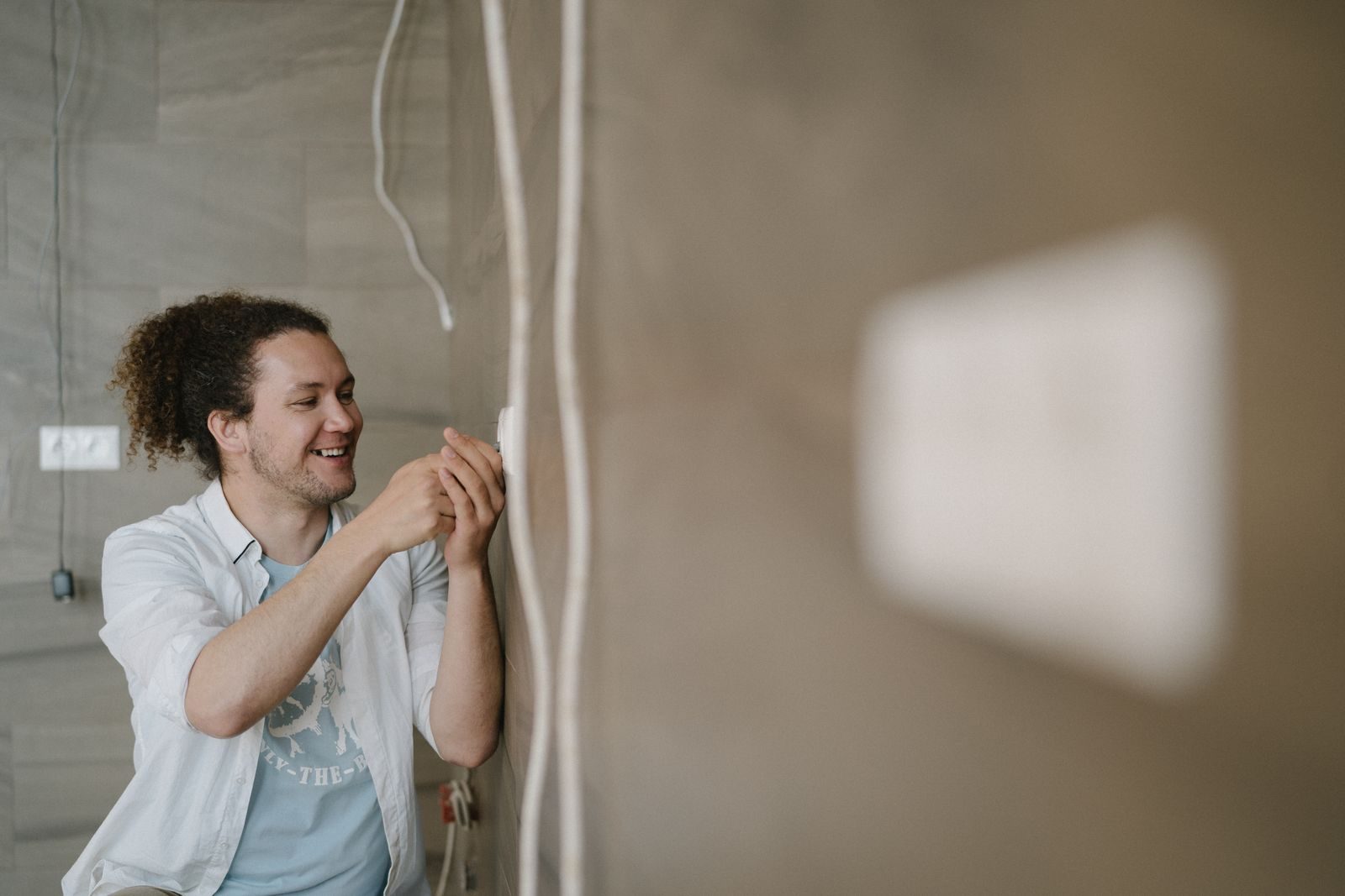 An electrician with curly hair adjusts an outlet indoors, smiling as he works
