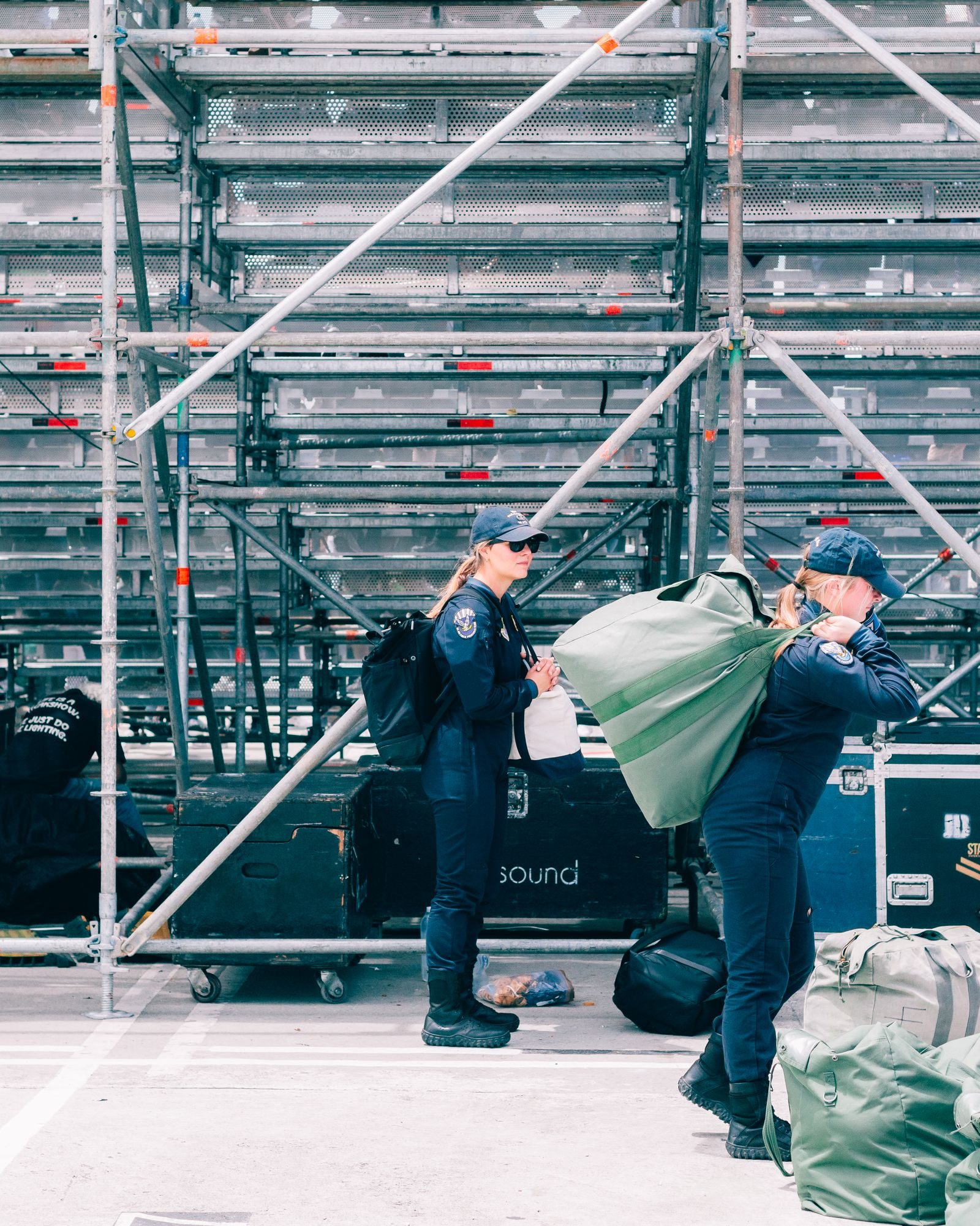 Two women in uniforms organizing bags and equipment outdoors against a metal backdrop