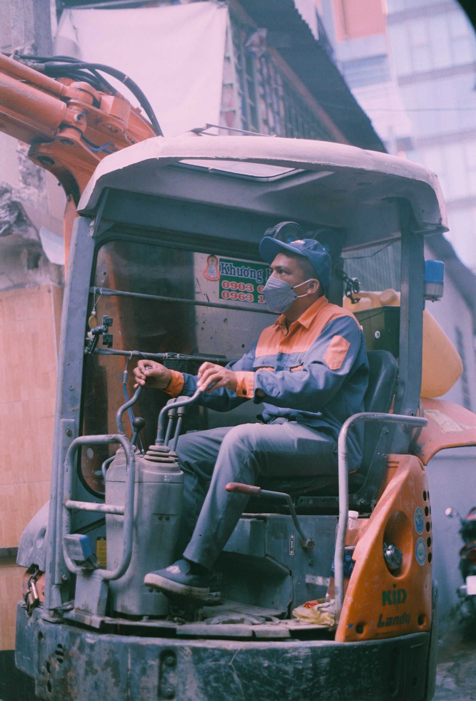 A male construction worker operates an excavator on a busy construction site, wearing a face mask and helmet