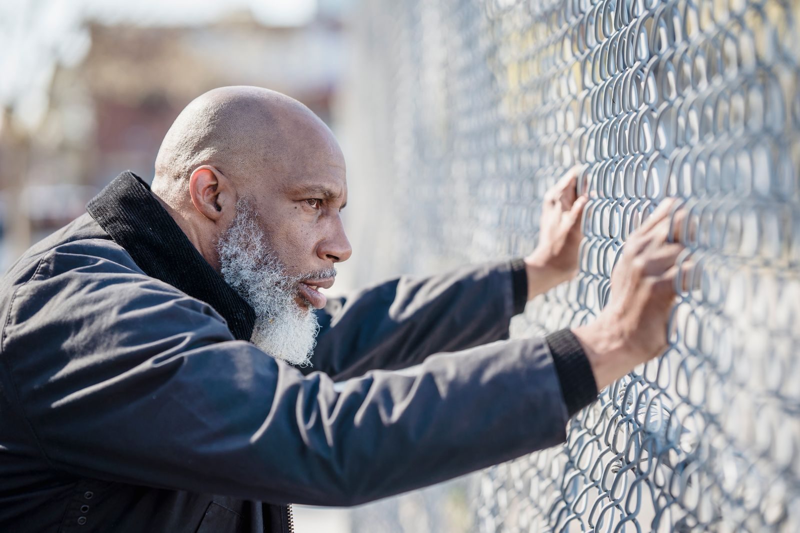 A bald man with a beard leans against a chain-link fence outdoors, looking thoughtful