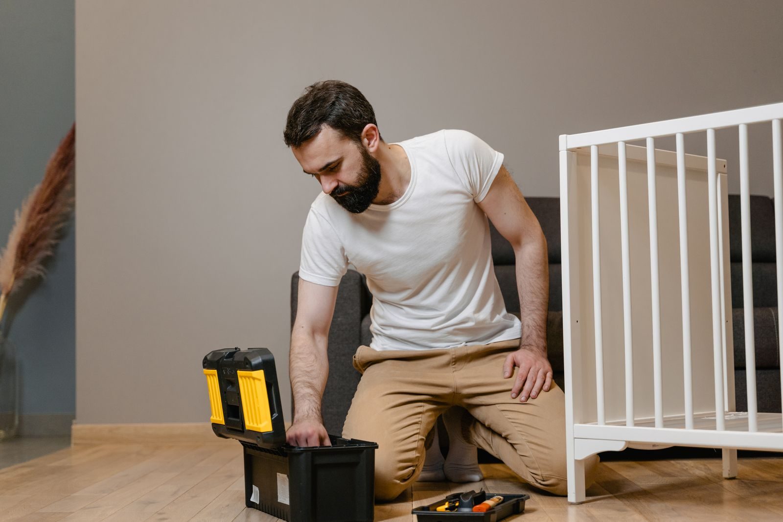 A man kneels indoors assembling a baby crib using a toolbox, wearing a white shirt and beige pants