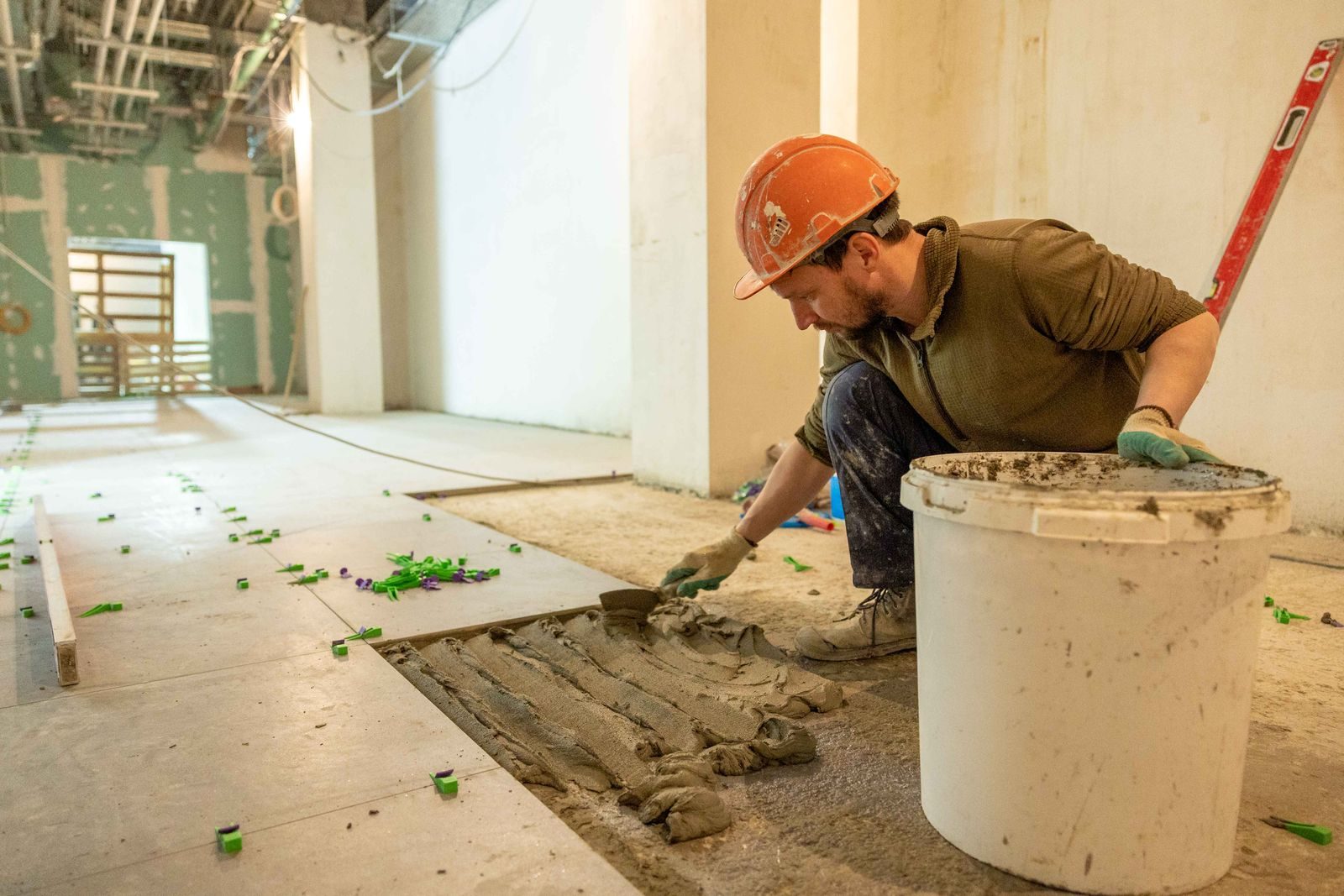 A construction worker lays tiles in an indoor renovation project, wearing safety gear