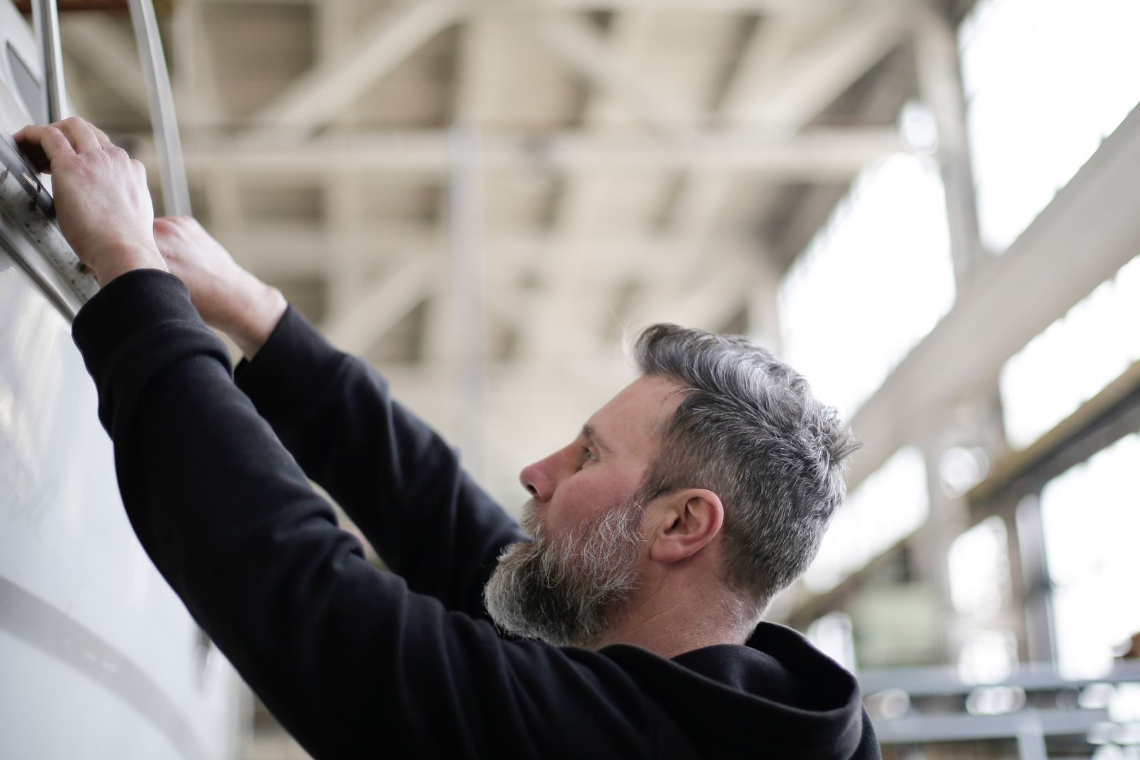 Side view of adult bearded mechanic in black workwear checking metal details while working in spacious modern