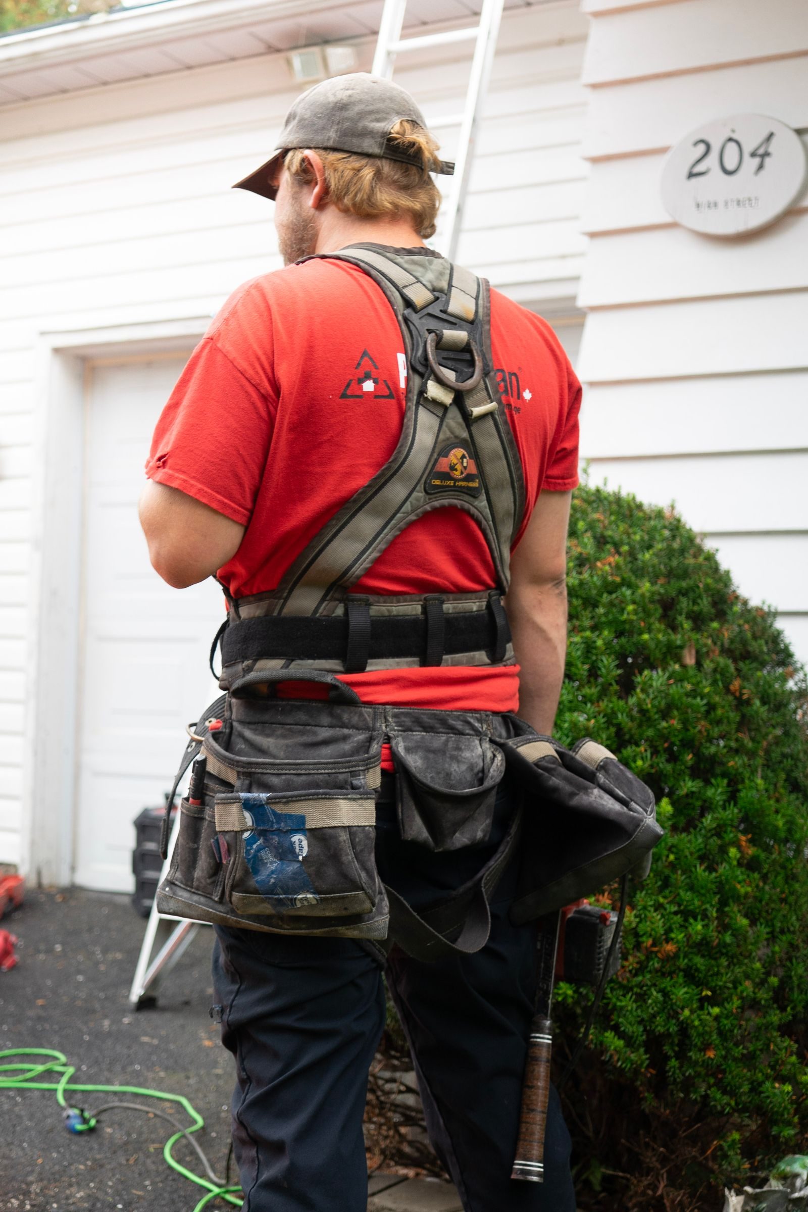 Back view of construction worker in red shirt and safety harness next to a house