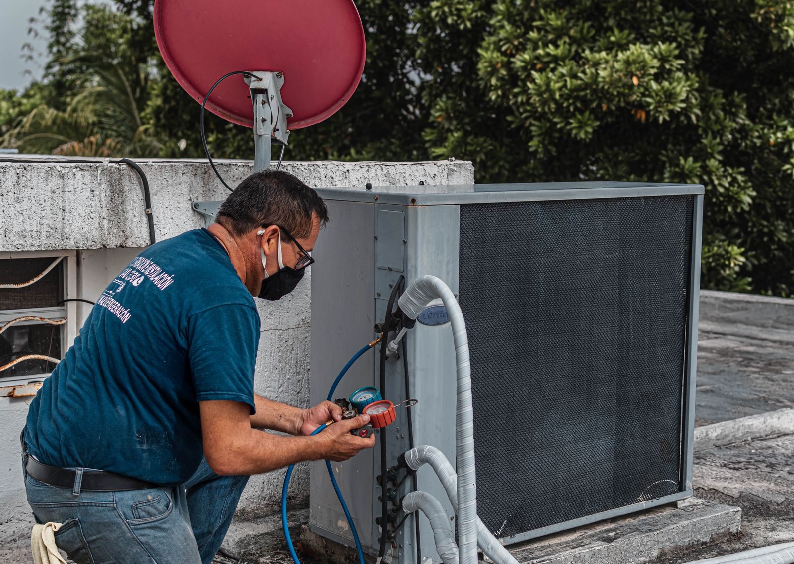 Technician repairing an air conditioner unit outdoors, wearing a facemask and using a manifold gauge