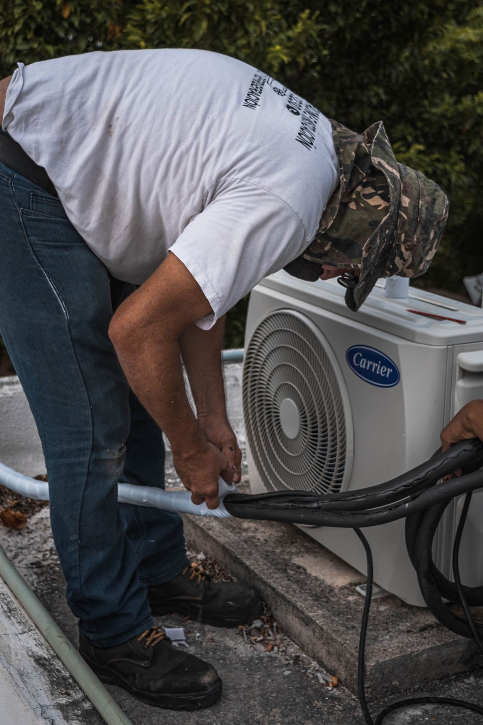 A technician performs maintenance on an outdoor air conditioning unit, focusing on hose connections