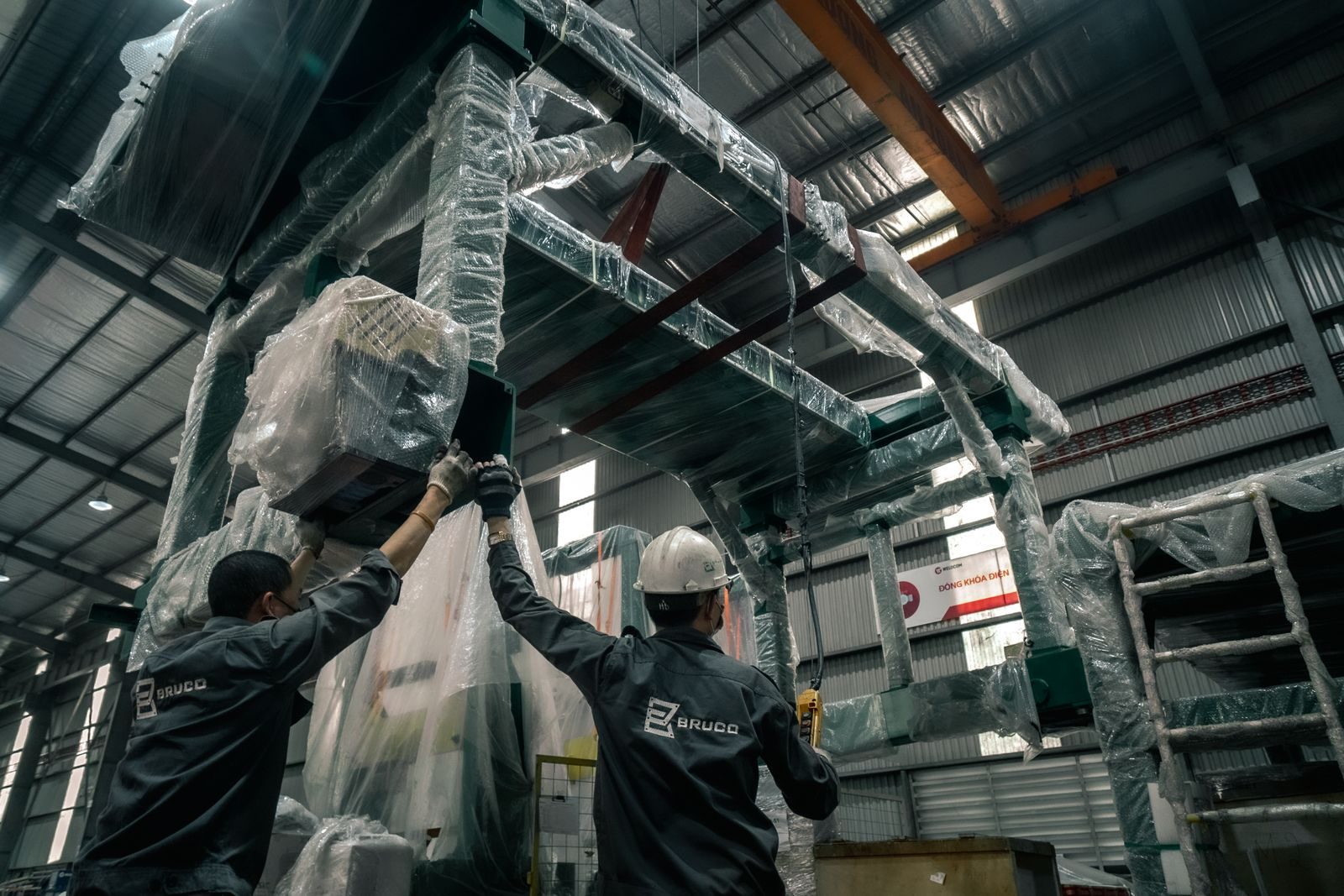 Two workers install large industrial machinery in a warehouse. They wear helmets and work with precision