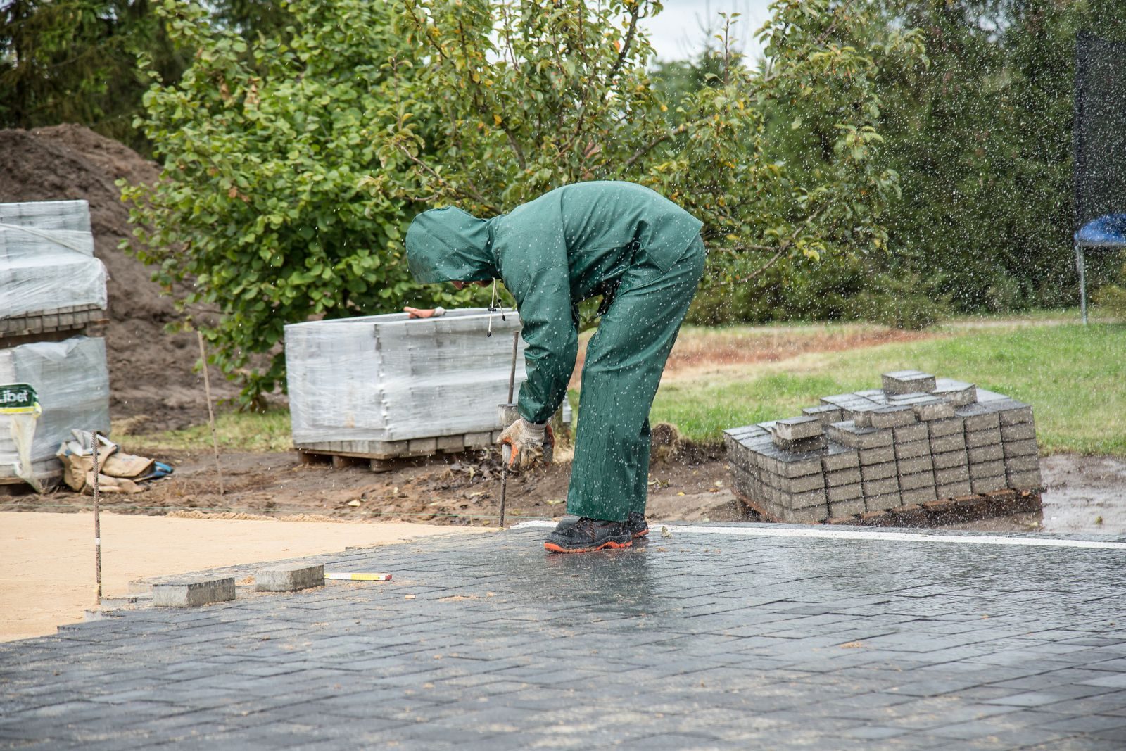 A worker in protective gear lays paving stones on a wet day