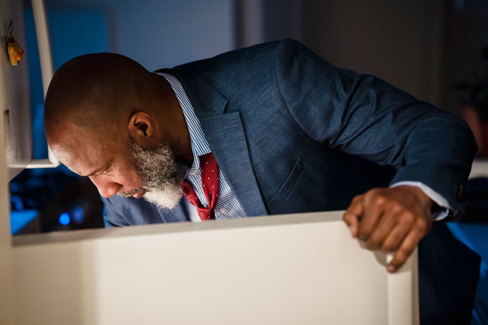 Smartly dressed man peering into a refrigerator, possibly late at night at home
