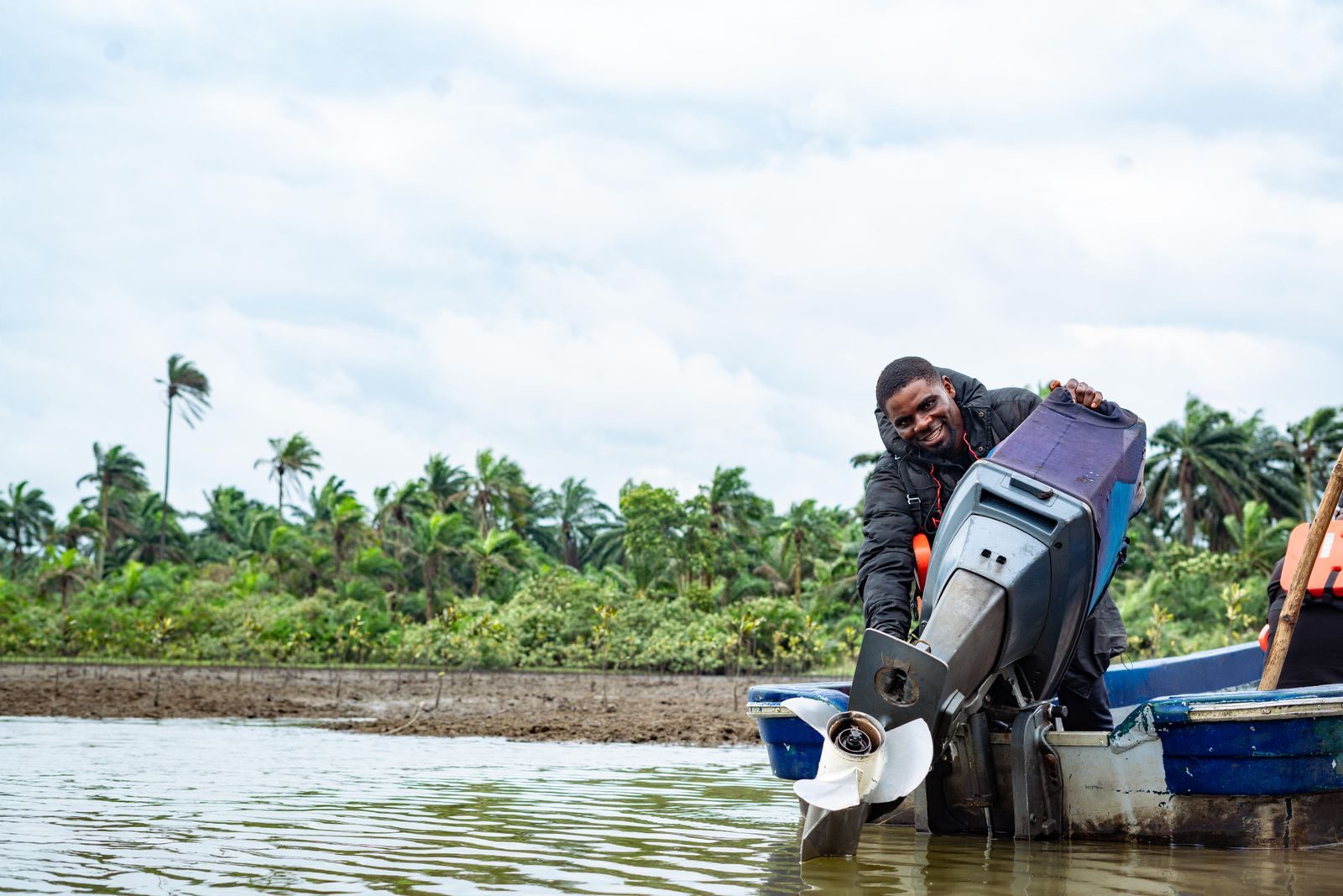 A man repairs a boat engine near tropical vegetation on a sunny day