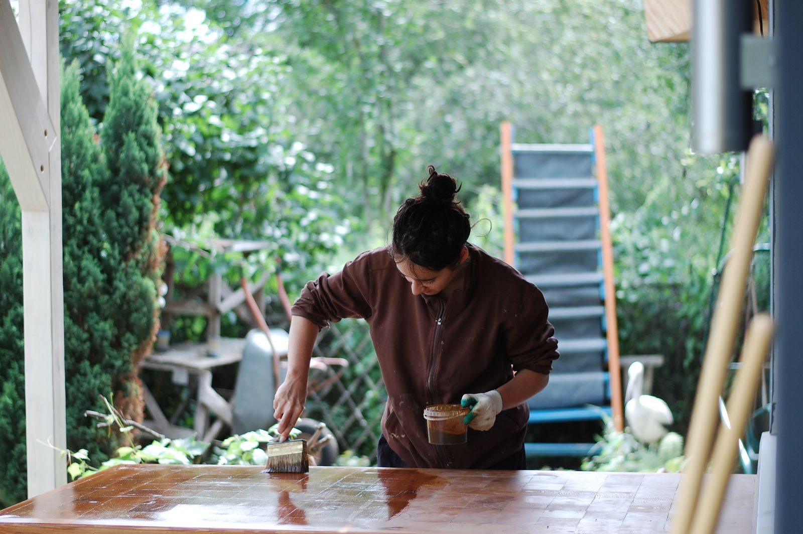 Woman applies varnish to a table outdoors, surrounded by greenery. DIY and home improvement scene