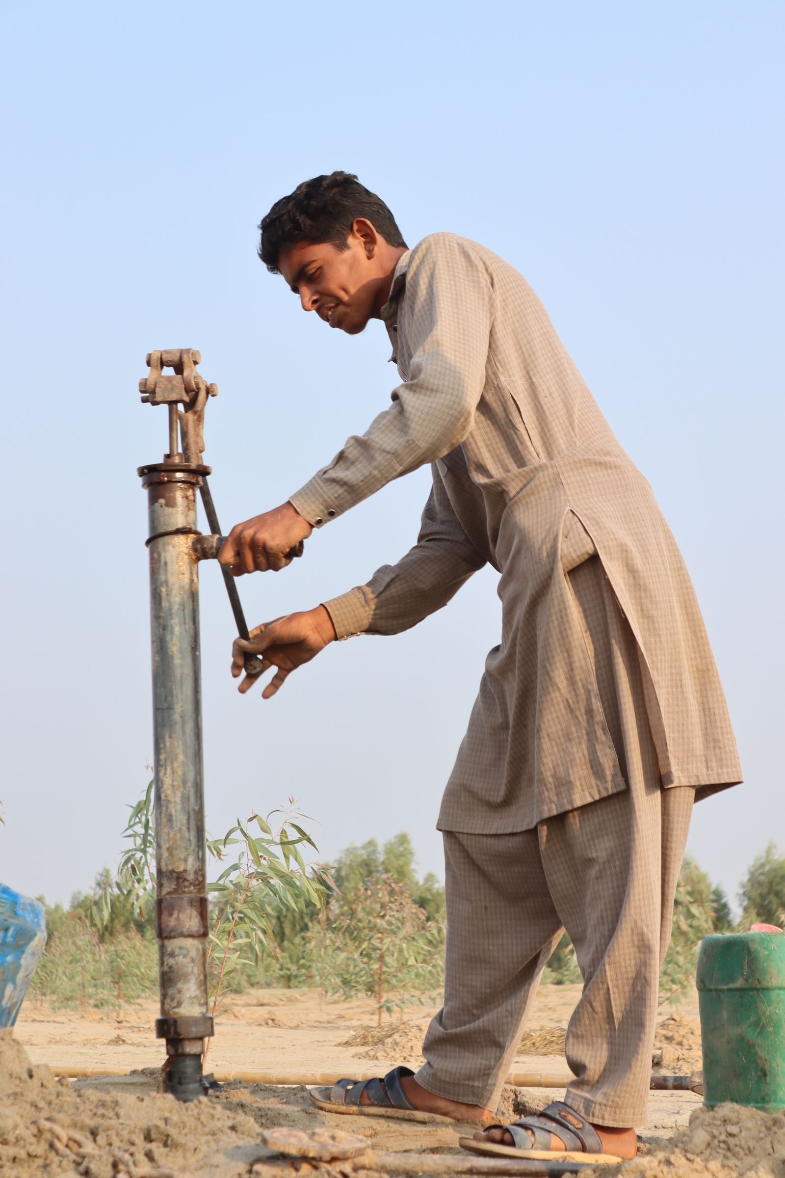 A man operates a hand pump in a rural setting, wearing traditional attire