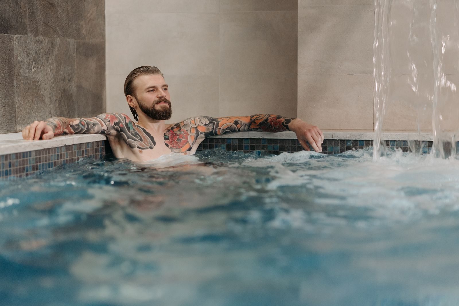 A shirtless tattooed man with a beard relaxing in an indoor pool with waterfall effects