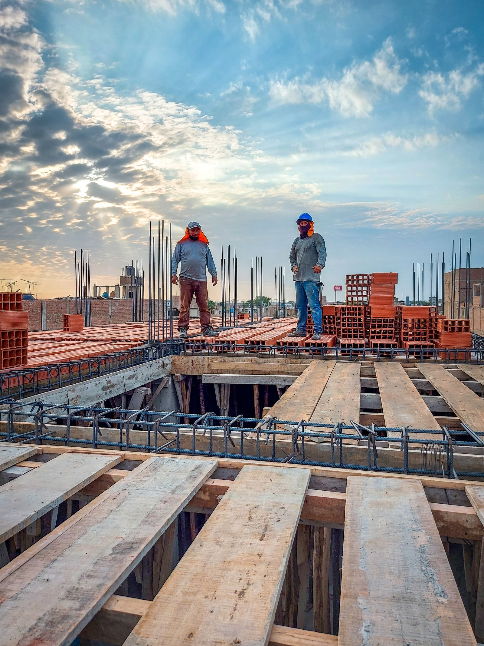 Two construction workers stand on a building site under a cloud-filled sky. Sunlight illuminates the scene