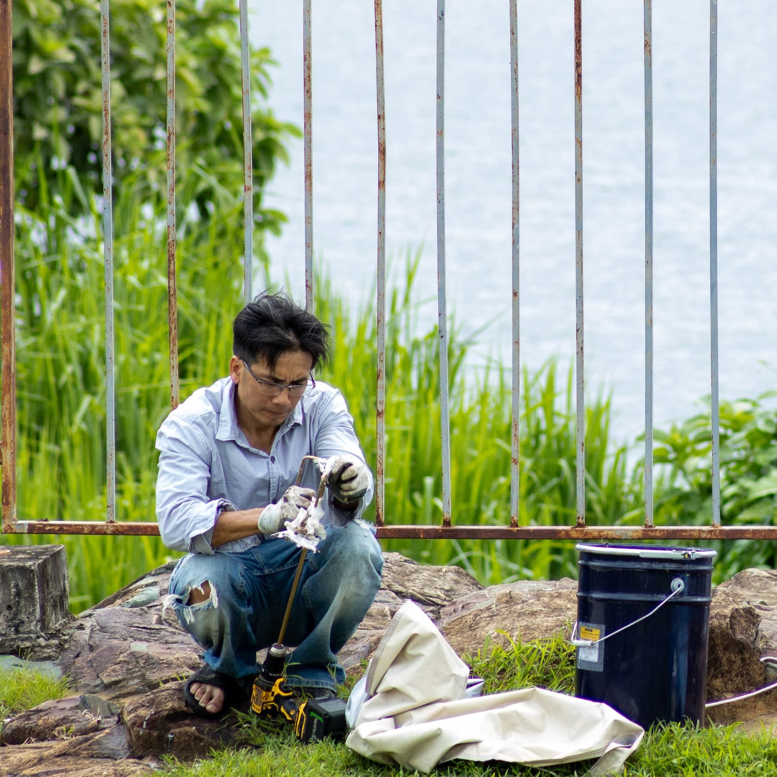 An adult male engaged in outdoor maintenance work by a lakeside, using tools and wearing gloves