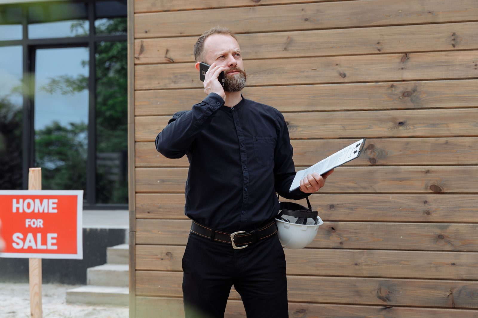 Real estate agent making a phone call while holding documents near home for sale sign