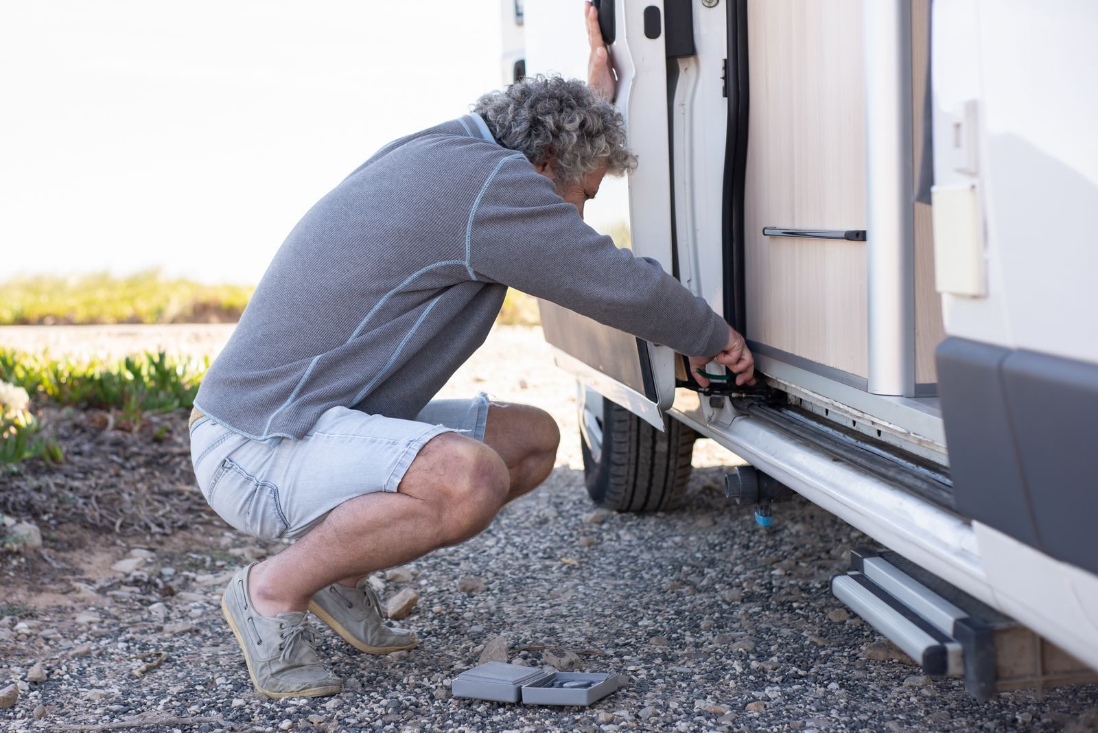 Senior man fixing camper van door mechanism outdoors. Emphasizes travel and hands-on problem-solving