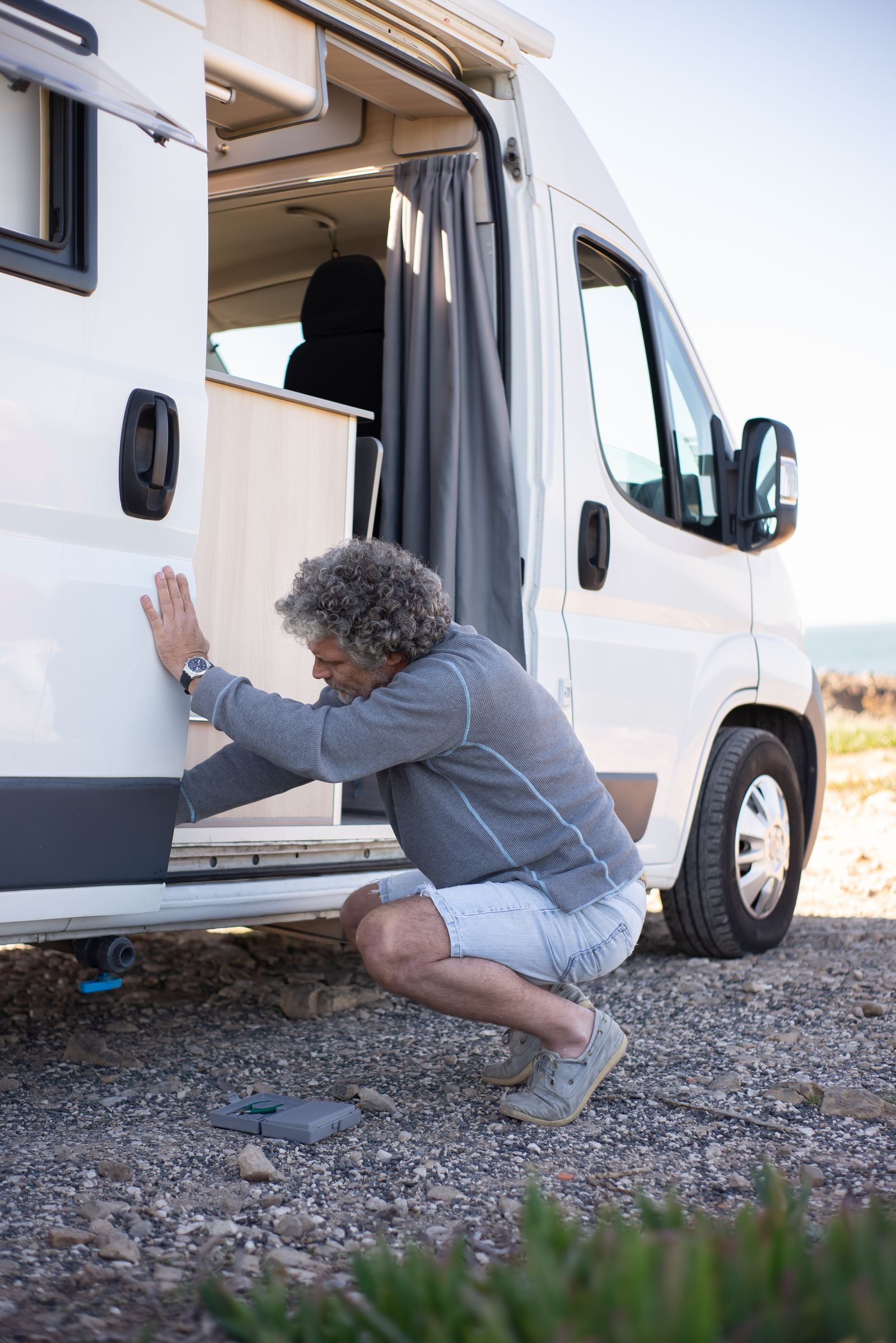 An elderly man in a gray sweater and shorts fixes his camper van outside, capturing the essence of travel and