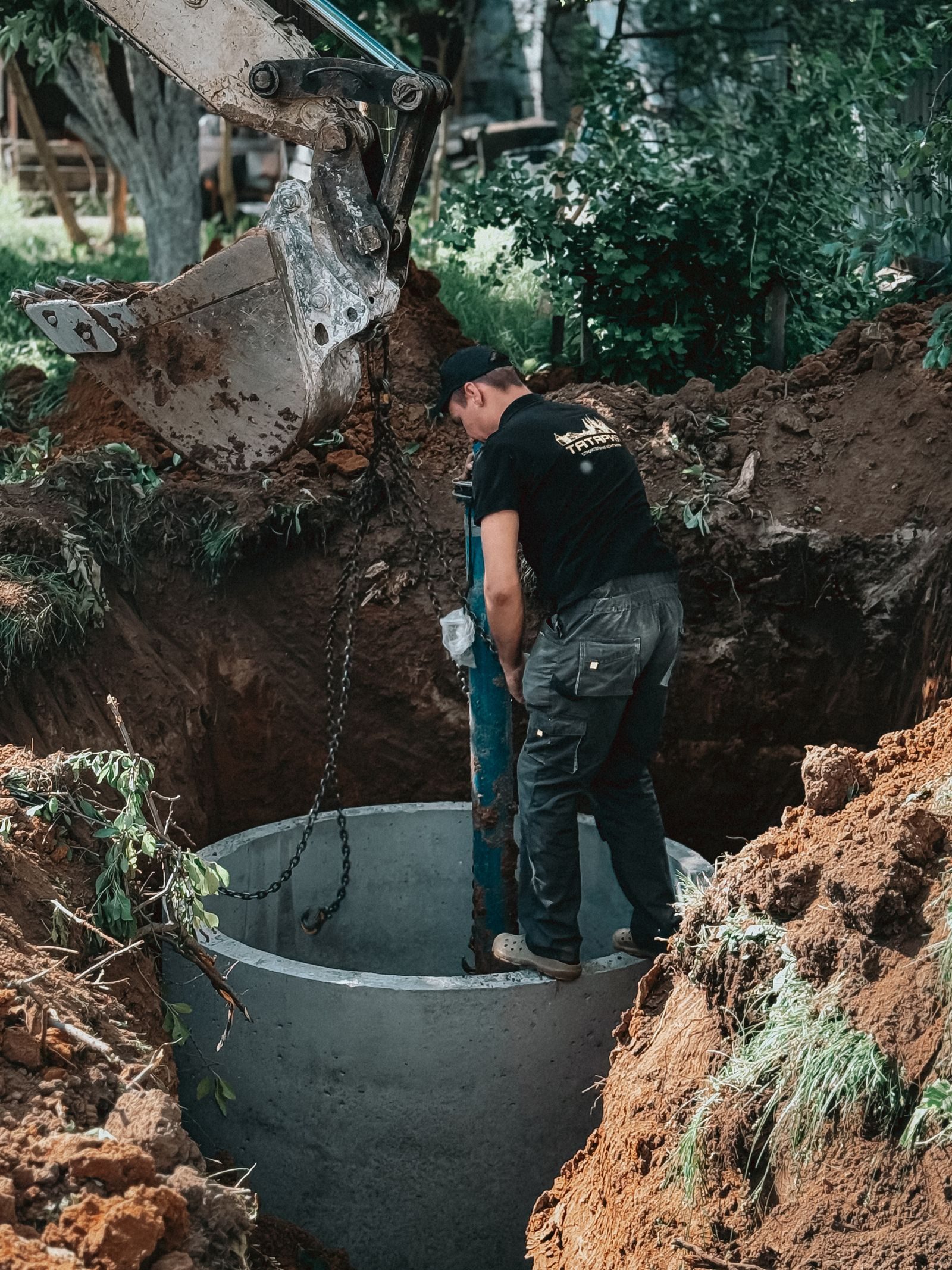 A construction worker operates heavy machinery to install a concrete pipe into the ground, surrounded by dug s