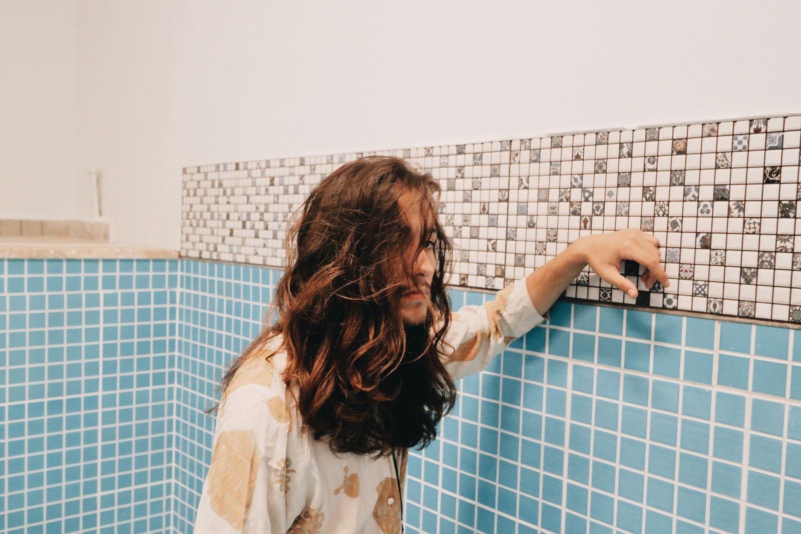 Young man with long hair posing indoors against a decorative tiled wall