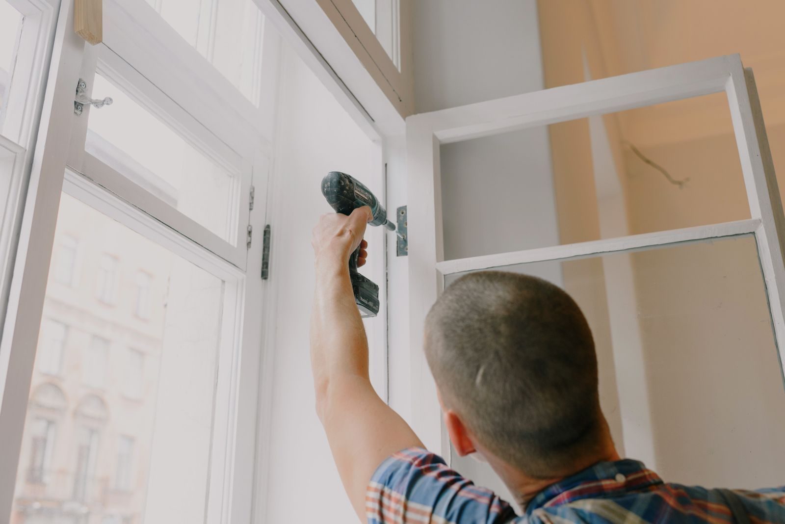 Back view of unrecognizable craftsman using cordless screwdriver and assembling window while screwing metal hi