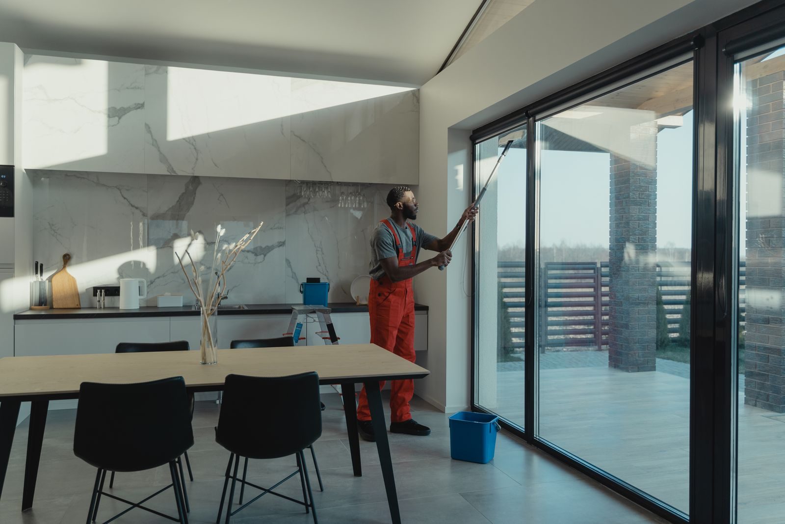 A professional cleaner in uniform washing large glass windows indoors with natural light streaming in